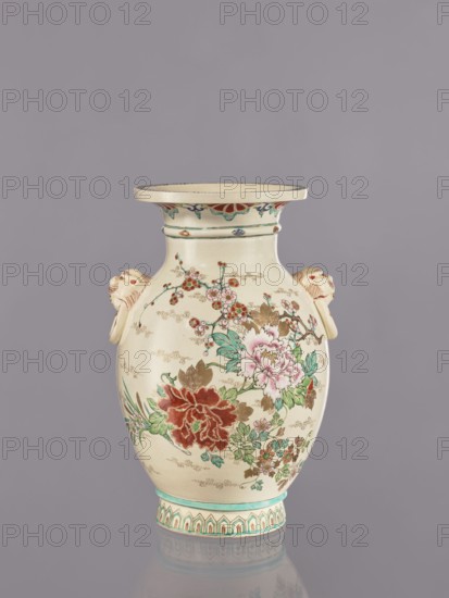 Vase, decorated with flowers and lions in satsuma ware; Japanese ceramics with cracklework decoration depicting peonies, chrysanthemums, and plum trees. Meiji period ca. 1870–1880. Vessel: height 26.5 cm; width 17.5 cm; diameter 16.5 cm., An off-white ceramic vase with a flared rim and a rounded body sits against a gray background; the vase features painted floral decorations in shades of red, pink, green, brown, and teal, with a large red flower and a pale pink flower amid green leaves and smaller blossoms, and delicate gold dotted accents scattered across the surface; there are sculpted handles on either side shaped like stylized animal heads in the same off-white with light brown shading, a decorative band with teal and brown motifs around the neck, and a patterned teal and brown border at the base, with the vase casting a faint reflection on the surface beneath it.