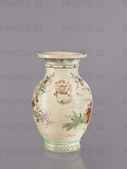 Vase, satsuma earthenware, decorated with crackle glaze and painted floral motifs including peonies, chrysanthemums, and plum trees, also featuring lions; Meiji period ca. 1870–1880, Japan; vessel; height 26.5 cm, width 17.5 cm, diameter 16.5 cm., An off-white ceramic vase with a flared rim and a rounded body sits against a muted gray background; the vase is decorated with hand-painted floral motifs in pink, red, green, and teal, with additional small brown and gold accent dots and fine brushwork; a sculpted lion-head ring handle in a similar off-white tone is attached to the upper body; the neck and base feature patterned bands in teal and beige with repeating geometric and foliate shapes; the vase casts a soft reflection on the surface beneath it.