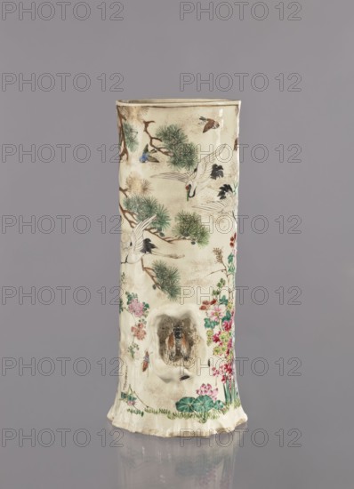 Vase, Tôritei maker, satsuma, Japanese, decorated with insects, tree trunks, flowers, plant ornaments, birds, peonies, pines, crickets, monkeys, kacho flowers and birds, kachoga iconography, lotuses, irises, grasses, cranes; Meiji period 1868–1912, ca. 1885 Meiji 18; Japan: Kyoto, Awata; ceramic vessel; height 32 cm, width 15 cm., A tall cylindrical ceramic object stands against a plain gray background, its surface predominantly off-white with a slightly aged, creamy tone. The object is decorated with painted natural motifs including green pine foliage, branches in brown, and several birds rendered with white, black, and touches of blue and brown; there are floral clusters in shades of pink, red, and green near the lower portion. A recessed hollow area on the front reveals a small sculpted figure with dark and orange-brown coloring set within a textured cavity. The base edge is irregular and shows some darker beige and brown variations, and the overall piece has fine speckled detailing...