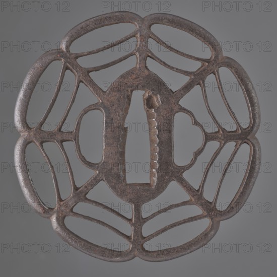 Crossguard tsuba, Tomotsugu; Japanese, Edo period 1603–1868, 19th century, Japan. Openwork tsuba Tsuba sukashi in the shape of a seven-petaled mokkô rosette, with openwork petals divided by arched lines forming a spiderweb pattern, integrally woven ryôhitsu ana holes and an oval seppa-dai. Signed. Materials: metal sword crossguard. Dimensions: height 8.0 cm, width 7.8 cm, depth 0.6 cm., A circular metal object with an openwork design resembling a symmetrical floral or web-like pattern, showing a central oval opening with a vertical rectangular slot and textured inner edges; the metal surface is dark brown with areas of reddish-brown and subtle black patina, and the background is a uniform gray.