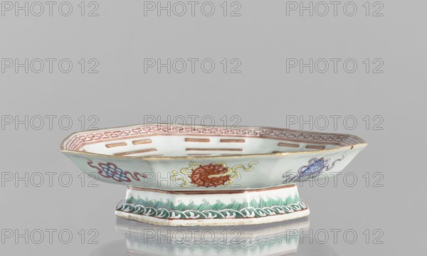 Platter, ceramics, colored porcelain, Qing dynasty Tongzhi period, 1862–1874, Chinese, diameter 24.5 cm, height 5.8 cm. In the mirror, the Taoist sign Taiji appears as two stylized intertwined birds, surrounded by the Eight Immortals and their corresponding trigrams. The outer rim is decorated with Buddhist symbols. Vessel., A shallow, footed porcelain dish with a pale greenish-white body sits against a neutral gray background; the dish has a gently flared rim with a gold-colored edge and a decorative red patterned band inside the rim, and the exterior shows a few painted motifs in red, yellow, blue, purple, and green, including a central circular motif with flowing ribbon-like elements and additional stylized designs on either side; the foot is decorated with green foliate and wave-like patterns and the dish casts a soft reflection on the surface beneath it.