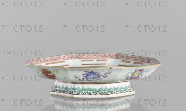 Platter;, Colored porcelain, Qing dynasty Tongzhi period, 1862–1874, China. Decorated in the mirror with the Taoist Taiji sign as two stylized birds intertwined, surrounded by the Eight Immortals and their corresponding trigrams; outer rim decorated with Buddhist symbols. Vessel; height 5.8 cm, diameter 24.5 cm., An octagonal shallow porcelain dish on a short pedestal, shown from the front against a gray background, with pale celadon green exterior and interior surfaces, a gold rim, and decorative motifs in red, purple, yellow, blue, green, and white; the interior rim has a repeated geometric border in red, the central interior shows several elongated cutouts, the exterior sides display a few stylized floral and ornamental emblems in red, purple, yellow, and blue with curling ribbon-like elements, and the pedestal bears a green wave-like painted band above a white base, with the dish casting a soft reflection on the surface below.