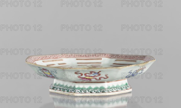 Platter, ceramics, colored porcelain, Qing dynasty, Tongzhi period 1862–1874, Chinese, In the mirror: the Taoist sign Taiji as two stylized intertwined birds, surrounded by the symbols of the Eight Immortals and their corresponding trigrams; the outer rim decorated with Buddhist symbols. Vessel; height 5.8 cm, diameter 24.5 cm., A shallow, octagonal ceramic dish with a raised pedestal base sits against a pale gray background. The dish surface is a soft pale green with painted decorative motifs in colors including red, pink, blue, yellow, green, and gold. A gold rim edges the top of the dish, and a red geometric border runs inside the rim. The exterior shows painted stylized floral or ribbon-like designs in red, blue, pink, and yellow, and the base has green leaf-like patterns with a darker green wave or braid motif along its lower edge. The interior has a series of elongated, rectangular cutouts and red linear decorations visible against the pale green field. The piece rests on a refle...