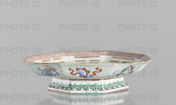 Platter, ceramics, colored porcelain, Qing dynasty, Tongzhi period 1862–1874, Chinese, decorated with the Taoist Taiji sign in the form of two stylized birds intertwined, surrounded by the Eight Immortals and their corresponding trigrams; outer rim decorated with Buddhist symbols. Vessel; height 5.8 cm, diameter 24.5 cm., A shallow, octagonal porcelain dish with a raised foot, displayed against a plain light gray background, showing pale celadon and white glaze with painted decorative motifs in colors including green, blue, red, brown, and gold; the rim has a reddish-brown border pattern, the exterior sides feature a few stylized floral and ribbon-like designs in blue and red with touches of yellow and brown, and the foot is decorated with green wave-like patterns and small pale dots; the dish casts a soft reflection on the glossy surface below.