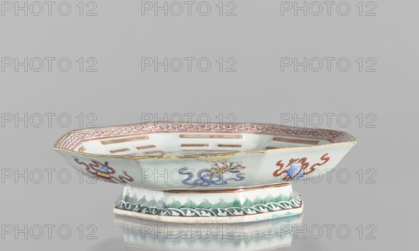 Platter, ceramics, colored porcelain, Chinese, Qing dynasty Tongzhi, 1862–1874. In the mirror, the Taoist sign Taiji appears as two stylized birds intertwined, surrounded by the symbols of the Eight Immortals and their corresponding trigrams. The outer rim is decorated with Buddhist symbols. Vessel; height 5.8 cm, diameter 24.5 cm., A low, shallow ceramic dish with an octagonal rim sits on a short octagonal foot; the dish is predominantly pale light green or celadon with painted decorative motifs in blue, red, green, yellow, and gold along its sides and inner rim, including floral and ribbon-like designs and a repeating geometric border inside; the exterior rim has a worn gold trim, and the foot shows green leaf-like patterns and a band of darker green on a white ground; the piece rests on a reflective grey surface against a plain light grey background.