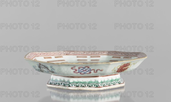Platter, ceramics, colored porcelain, painted with the Taoist Taiji symbol as two stylized intertwined birds, surrounded by the Eight Immortals and their corresponding trigrams; outer rim decorated with Buddhist symbols. Qing dynasty, Tongzhi period 1862–1874, China. Vessel. Height 5.8 cm; diameter 24.5 cm., A shallow, footed ceramic dish with an octagonal rim sits centered on a reflective surface; the dish is primarily pale celadon with painted decoration in red, blue, green, yellow, and gold, including interlaced knot motifs and stylized cloud or flame elements around the exterior and a red patterned border along the inner rim; the foot is decorated with green leafy or wave-like patterns and small circular dots, and the rim has a thin gold edging; the background is a uniform light gray.