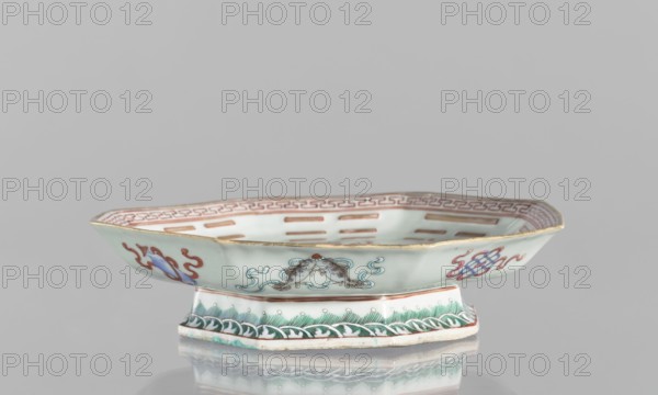 Platter, colored porcelain; Qing dynasty, Tongzhi period 1862–1874; China. Circular vessel, height 5.8 cm, diameter 24.5 cm. Decor: central mirror with the Taoist Taiji sign rendered as two stylized birds intertwined, surrounded by the Eight Immortals with their corresponding trigrams; outer rim decorated with Buddhist symbols., A shallow octagonal ceramic dish on a matching pedestal base, shown from the front on a reflective surface, with a pale celadon body and a gilt rim. The exterior features painted motifs in red, blue, turquoise, brown, and green, including a pair of stylized animal or fish figures with flowing turquoise lines, curving red decorative elements, and a linked knot-like design in blue and red. The pedestal has a band of green leaf-like patterns above a border of white and green wave motifs, and the interior rim shows a repeated geometric border in red with pale celadon on the dish floor.