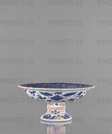 Platter, Narozno Eckersdorf, phoenixes iconography, ceramics, platters, Japanese art, Japanese culture, floral ornaments, colored porcelain, gardens, birds, Edo 1603–1868, early 18th century, Japan, Arita, vessel, height 9.7 cm, diameter 21.7 cm, A shallow pedestal dish sits centered against a plain gray background, showing white ceramic decorated with bold blue floral and foliate motifs and additional orange-red accents; the wide rim flares outward and the interior shows blue panels with orange-red detailing, the exterior of the bowl displays flowing blue leaves and scrolls punctuated by orange-red curls, and the short cylindrical stem and round foot are similarly ornamented with bands of blue leaves and orange-red decorative patterns, with a subtle reflection of the piece visible on the surface below.