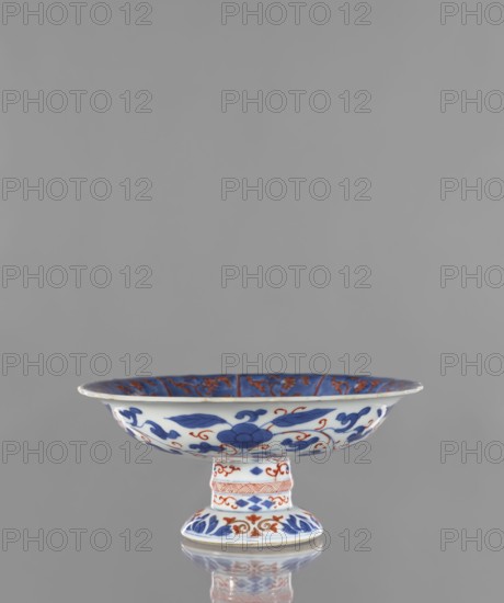 Platter, Narozno Eckersdorf; ceramics, colored porcelain; Arita, Japan; early 18th century Edo, 1603–1868; platter/vessel, height 9.7 cm, diameter 21.7 cm; decorated with phoenixes, birds, floral ornaments, gardens; Japanese art/culture; provenance notes: repossession., A shallow pedestal bowl sits centered against a plain gray background, the bowl and foot decorated with floral and foliate motifs in blue and orange on a white ground; the bowl interior shows a deep blue rim with orange floral patterns, the exterior features blue flowers and leaves with orange scrolling accents, and the pedestal foot displays alternating blue and orange decorative bands and motifs, the piece shown with a subtle reflection beneath it.