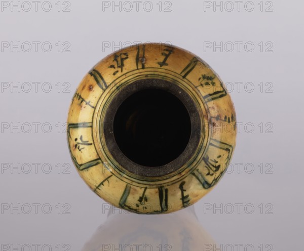 Vase, repossession Glogów, Islamic art, Persian culture, glazed ceramic utility vase, lambrequin pattern, 18th century, Iran, vessel; height 31.5 cm, diameter 19.7 cm, A round ceramic vessel viewed from above with a dark circular opening at its center and a yellowish-beige body decorated with green painted rectangular panels and short vertical strokes; the rim shows a darker brownish inner edge and the surface has small brown spots and areas of wear; the background is a pale gray and the vessel casts a faint reflection below.