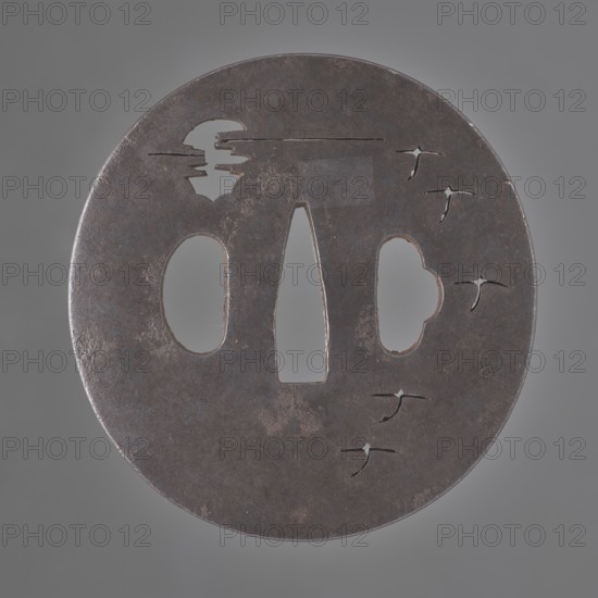 Crossguard tsuba, Tetsugendô,  school author, Japanese art, Japanese culture, Edo 1603–1868, weapons, cold weapons, swords, katana, sword crossguards, tsubas, moons, birds, geese, Tsuba round maru gata, flat, with a polished surface migaki ji. Decorated with a delicate negative openwork creating a motif of a moon partially hidden behind clouds and a flight of wild geese. Unsigned. 18th century Edo period 1603–1868, Japan, tsuba, height 7.1 cm, width 6.9 cm, depth 0.35 cm, A round dark metal object with a worn, rusty surface sits against a neutral gray background; the object has a few cutouts including a central vertical elongated cutout and two irregular oval cutouts flanking it, a few smaller star-like slits near one edge, and an irregular silhouette cutout near the top, showing colors of dark brown, black, gray, and hints of reddish rust.