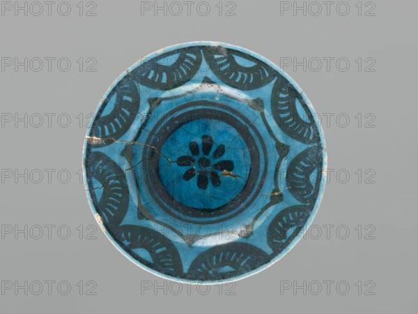 Plate, Islamic art, Persian culture, ceramics, plate/saucer, Caucasus, geometric motifs, decoration: black painted semicircles, stylized flower in the mirror, 17th century, Azerbaijan, vessel, height 2.0 cm, diameter 12.5 cm, A round ceramic plate with a glazed surface in shades of blue and dark teal, featuring a central floral-like motif surrounded by concentric circular bands and a repeating scalloped decorative pattern along the rim; the glaze shows visible crack lines and some small chipped or worn areas, and the background behind the plate is a flat neutral gray.