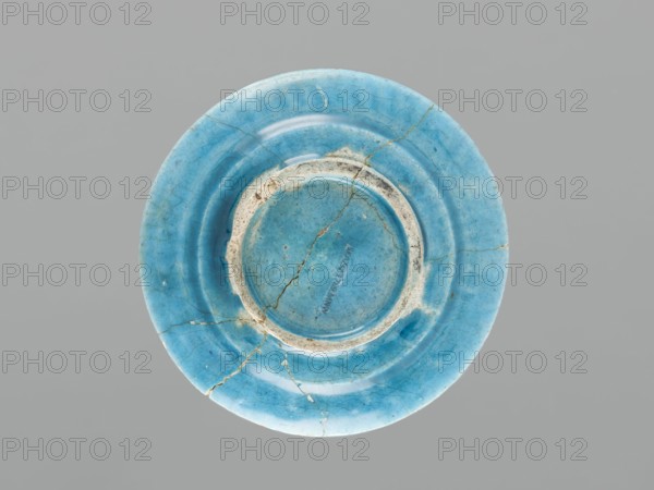 Plate, Islamic art, Persian culture, ceramics, plate/saucer, Caucasus, geometric motifs; decoration: black painted semicircles, stylized flower in the mirror; 17th century; Azerbaijan place of origin; vessel; height 2.0 cm; diameter 12.5 cm, A circular ceramic plate viewed from above with a smooth glossy surface in shades of blue and turquoise, showing a central recessed area and concentric raised rings; the plate has visible hairline cracks and a few larger repaired fractures with irregular light beige or off-white areas where the glaze is worn or chipped, and faint darker blue speckling across the surface; the background is a uniform neutral gray.