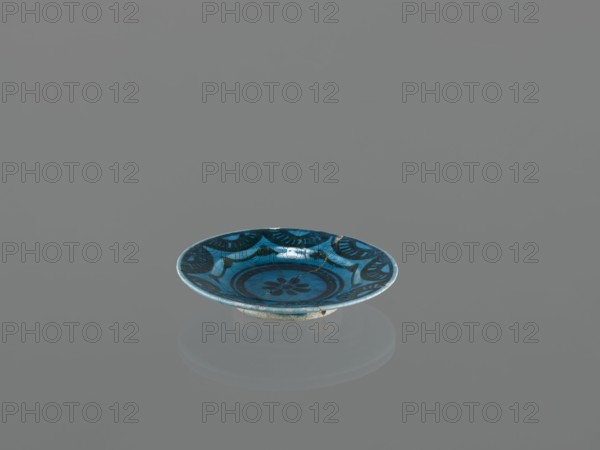 Plate, Islamic art, Persian culture, ceramic plate/saucer, Caucasus, geometric motifs; decoration: black-painted semicircles, stylized flower in the mirror; 17th century; Azerbaijan place of origin; vessel; height 2.0 cm, diameter 12.5 cm, A shallow round dish with a raised foot, predominantly blue with darker blue decorative patterns and lighter blue highlights, sits centered on a smooth grey surface; the dish shows a floral motif in the center and curved decorative bands toward the rim, and a soft reflection of the dish appears on the surface beneath it.