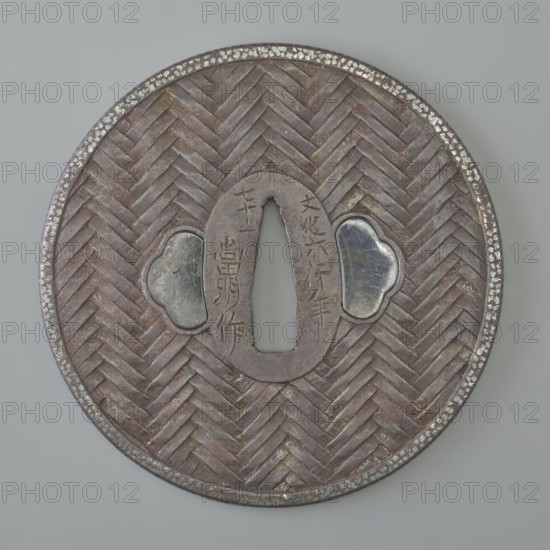 Crossguard tsuba, maker: Tômei, unknown provenance; Japanese Edo period, Bunka era 1809. Iron tsuba round maru-gata with rectangular edge kaku mimi; seppa-dai oval, ryohitsu ana blinded with shakudo. Both sides decorated in relief: braided motif—herringbone on the obverse, squares on the reverse. Edge with delicate ornament of densely arranged spots. Signed. Diameter 8.35 cm; thickness 0.4 cm., A circular metal object with a woven chevron pattern across its surface in shades of brown and dark gray, featuring a central elongated oval opening flanked by two cloud-shaped insets of shiny silver-gray, a raised rounded rim with patches of lighter silver, and an overall aged, slightly weathered appearance with subtle reddish-brown tones and darker worn areas.