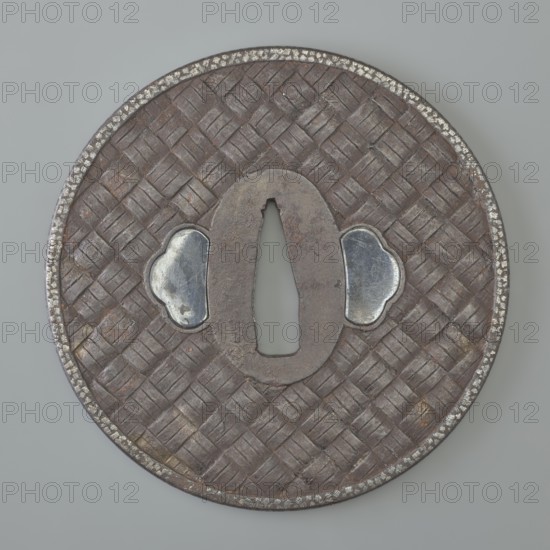 Crossguard tsuba, Tômei, sword crossguard from Japan, Edo period, Bunka era 1809. Tsuba round maru gata with rectangular edge kaku mimi; seppa-dai oval, ryôhitsu ana blinded with shakudo. Entire surface decorated on both sides in relief: braided motif—herringbone on the obverse, squares on the reverse. Edge decorated with a delicate ornament of densely arranged spots. Signed. Diameter 8.35 cm; thickness 0.4 cm., A round object with a woven textured surface in brown tones and a darker brown central oval opening, bordered by a thin rim that shows speckled silver wear, with two irregular silver-colored metal insets flanking the central opening, all set against a light grey background.
