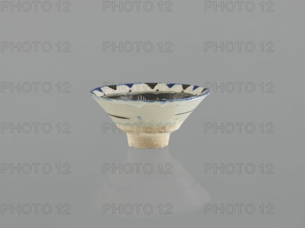 Bowl, Islamic art, Arabic culture, Ayyubid dynasty 1171–1341, ceramics, Arabic ceramics, Middle Eastern ceramics. Bowl on a ring-shaped foot, wide bottom, inverted-cone shape characteristic of Syrian bowls and goblets produced during the Ayyubid period. Likely quartz ceramic mass probably made in Egypt in the 12th century, a feature of Ayyubid wares. Decoration: stylized plant motifs — a lotus flower and a leaf — in the mirror, surrounded by rays of light; linear and radial ornament painted in black; stylized flower in the mirror. Decoration executed in a free brush style popularized by Syrian artists in the 12th–13th centuries; composition shows formal simplification typical of contemporary Middle Eastern ornamentation. M.P., 13th century, Syria; vessel; weight 0.26 kg; height 7.4 cm; diameter 15.9 cm., A small shallow ceramic bowl sits centered on a smooth reflective surface, showing a muted reflection below. The bowl has an off-white to cream base color with a slightly rough unglaze...