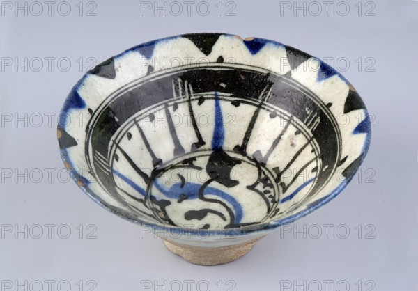 Bowl, Islamic art, Arabic, Ayyubid dynasty 1171–1341, ceramics, Arabic ceramics, Middle Eastern ceramics. Bowl on a ring-shaped foot with wide bottom and inverted-cone profile. Quartz ceramic body probably Egypt, 12th century. Decorated in black with linear and radial ornament: stylized plant motifs—a lotus flower and a leaf—surrounded by rays of light; stylized flower in the mirror. Decoration painted in a free brush style associated with Syrian artists of the 12th–13th centuries; composition shows formal simplification characteristic of the period. 13th century, Syria, vessel; weight 0.26 kg; height 7.4 cm; diameter 15.9 cm., A shallow ceramic bowl seen from above showing its interior and rim, with a few areas of glaze missing on the edge revealing the underlying clay; the bowl has a white base color with several decorative black brushstroke patterns and a few bold blue brushstrokes forming radial and curved motifs; the rim features a repeating triangular pattern in black and blue ag...