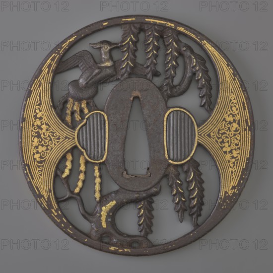 Crossguard tsuba, Japanese, Edo period 1603–1868, early 18th century, iron tsuba, maru gata round, kaku mimi rectangular edge, ryôhitsu-ana oval blinded with shakudô. Decorated in yô sukashi positive openwork with a hoo phoenix perched on a willow tree; decoration and edge with gold inlay using the nunome technique. Aizu-Shôami style. Not signed. Height 8.4 cm; width 8.2 cm; depth 0.45 cm., A round ornamental metal object with a central elongated opening, showing a dark brown patina and areas of worn gold decoration; the outer rim is circular and dark with scattered gold highlights; on one side a stylized bird with spread wings is visible in dark brown with touches of gold on its beak and body; hanging leafy or fern-like forms in dark brown descend near the top and sides; two oval ridged forms flank the central opening, each outlined with gold and showing dark ridged interiors; sweeping gold filigree patterns decorate two symmetrical curved panels on opposite sides, with fine gold line...