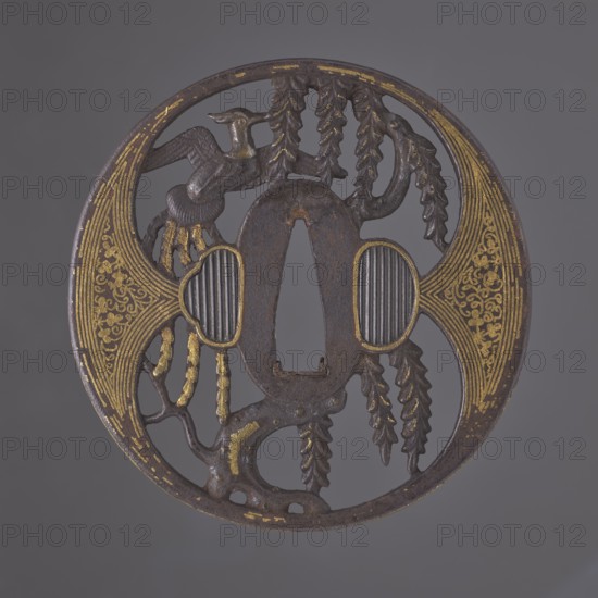 Crossguard tsuba, Japanese, Edo period 1603–1868, early 18th century, weapons — katana sword crossguard tsuba, maru gata round, rectangular edge kaku mimi, oval ryôhitsu-ana blinded with shakudo. Decorated with a phoenix hoo perched on a willow tree cut out in positive openwork yô sukashi; decoration and edge have gold inlay using the nunome technique. Aizu-Shoami school; not signed. Dimensions: height 8.4 cm, width 8.2 cm, depth 0.45 cm., A round metal object with an openwork design showing a central oval opening and a few curved cutouts, decorated with a group of stylized organic shapes resembling branches and leaves and a bird-like form; the surface is dark brown with areas of worn gold-colored detailing and a few grayish raised ridged sections, all set against a plain gray background.