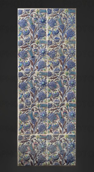 Panel of 10 tiles with plant decoration, Islamic art, Persian, Qajar dynasty 1785–1925, ceramics, plant ornaments, floral motifs tulips, carnations, 18th–19th century, Iran, height 172.0 cm, width 64.0 cm, A vertical rectangular panel composed of a grid of decorative square tiles, each tile featuring a repeating floral pattern with stylized blooms, buds, leaves, and curving stems. The dominant colors are deep blue and cobalt in the large flower shapes, complemented by lighter blues and turquoise in smaller blossoms and striped petal elements, with accents of pale green and yellow-green on some buds and leaves. There are brownish-maroon leaves and stems woven through the design, and soft cream or off-white serves as the background color of the tiles. The tiles are arranged in several horizontal rows, and thin grout lines separate them, creating a slightly segmented look across the overall patterned surface. The panel sits against a dark neutral backdrop that contrasts with the lighter t...