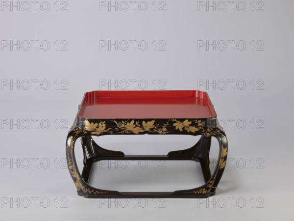 Tray on legs kakeban, noted. Japanese, Edo period early 19th century. One of a set of three tables placed one on top of the other; almost square red tops with high edges and bow legs connected at the bottom by a square frame forming the base. Sides and legs covered with black lacquer and sprinkled with gold using the maki-e technique with flowering cherry plum? and pine branches, depicting plum trees, flowers, trees, pines, twigs, winter. Piece of furniture. Height 25.3 cm; width 47.5 cm; depth 46.0 cm., A low rectangular tray table with a glossy red top set into a black frame, the black legs and apron decorated with gold floral and vine motifs, the legs curving inward to form open rectangular supports, the overall color palette including red, black, and gold against a light gray background.