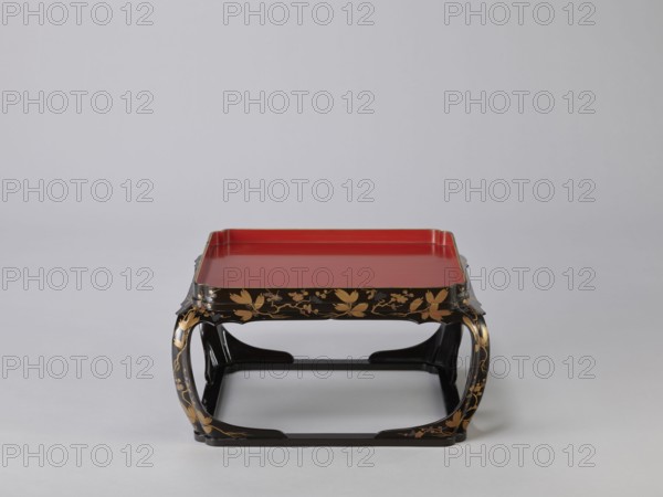 Tray on legs kakeban, One of a set of three tables placed one on top of the other; almost square red tops with high edges and bow legs connected at the bottom by a square frame, forming the base of the table. Sides and legs covered with black lacquer and decorated with gold powder using the maki-e technique in a motif of blooming plum and pine branches; motifs include plum trees, flowers, trees, pines, twigs, winter. Early 19th century Edo period, Japan. Piece of furniture. Height 22.0 cm; width 41.5 cm; depth 40.0 cm., A low, square tray table with a red flat top and raised edges sits centered on a light gray background, supported by a black lacquered frame with curved legs. The black frame is decorated with gold floral and leaf motifs along its sides and legs. The red surface contrasts with the black and gold of the base, and the overall scene shows muted shades of gray in the surrounding area.