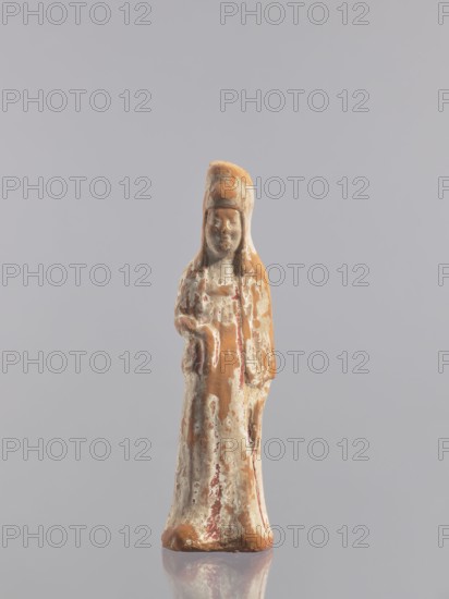 Standing woman figure - tomb figurine, Chinese, ceramics, female figurine, Tang dynasty 618–907, early 10th century, China, sculpture, height 19.3 cm, A small weathered figurine stands upright against a plain light gray background, showing a long robe and a tall rounded headdress; the surface of the figure is a mix of earthy brown, off-white, and muted reddish tones with areas of worn texture and flaking, and the base reflects faintly on the smooth surface beneath it.