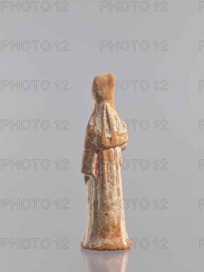 Standing woman figure - tomb figurine, unknown creator/craftsman, Chinese, ceramics, female figurine, Tang dynasty 618–907, early 10th century, China, sculpture, height 19.3 cm, A small worn figurine standing upright on a reflective surface, with an elongated robe-like shape and a hood or head covering; the surface shows patches of pale beige, terracotta brown, off-white, and faded red, with areas of flaking and uneven texture, and the background is a smooth light gray.