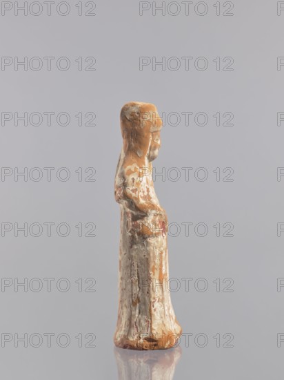Standing woman figure — tomb figurine; creator, Chinese; ceramics; female figurine; Tang dynasty 618–907, early 10th century; China; sculpture; height 19.3 cm, A small standing figurine with a weathered surface in shades of brown, beige, off-white, and hints of muted red, shown in profile against a plain light gray background; the figure appears to wear a long flowing garment and has a rounded head and simplified facial features, with areas of flaking and uneven texture exposing multiple layers of color, and the base of the object rests on a reflective surface that creates a subtle mirrored outline.