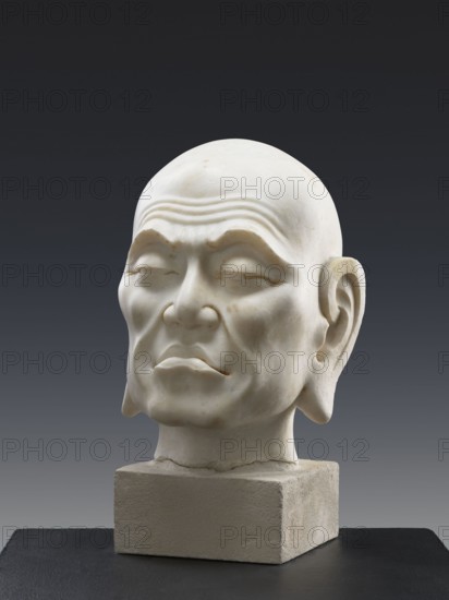 Head of Luohan, unknown sculptor, Chinese art, Chinese, Qing 1644–1911, Chinese Buddhism, arhat, monks, Luohan; according to information, China, 9th century. According to a Chinese specialist in Buddhist art, the sculpture appears to be contemporary and may come from the late Republic period, 19th century, China; sculpture, height 52 cm, A white stone or marble sculpted head of an older person with closed eyes and prominent brow lines sits on a textured rectangular base; the face shows pronounced cheekbones, a slightly downturned mouth, large ears, and subtle surface discolorations, all set against a dark gray to black gradient background with a black surface beneath the base.