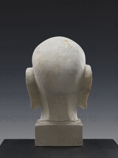 Head of a Luohan arhat, sculpture, Chinese; materials and, Iconography: Luohan/monk Chinese Buddhism. Dating uncertain: described as possibly 9th century but also suggested by a specialist to be contemporary, perhaps late Republic period, 19th century. Period: Qing 1644–1911 noted. Height: 52 cm., A rear view of a pale off-white stone or marble head sculpture with smooth rounded contours, visible ears that project outward, and a tapered neck set on a rectangular off-white base, all placed on a dark surface against a gradient dark-to-medium gray background, with subtle yellowish and gray discolorations on the stone.