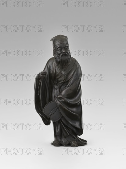 Sage, unknown sculptor, figurine of a Chinese sage wearing a cap and a long, flowing robe; depicts a man scholar. Chinese, Qing dynasty 1644–1911, 19th century. Sculpture, height 28.0 cm. Materials not specified., A dark brown to nearly black carved figure of an elderly man wearing flowing robes and a cap, standing on a light gray to white background; he has a long beard and mustache, a detailed face with a slight smile, one hand raised near his chest holding the edge of his robe and the other hand holding a rounded object; the robes show deep folds and drapery, and the figure stands on a small base with subtle carved feet and textured details.