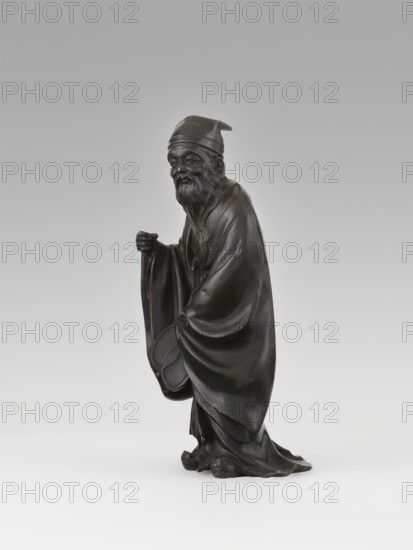 Sage, unknown sculptor; figurine of a Chinese sage man, scholar wearing a long flowing robe and a cap; fan; Qing dynasty 1644–1911, 19th century; China; sculpture; height 28.0 cm; unknown provenance., A dark bronze-colored statue of an elderly man wearing a long flowing robe and a soft hat, standing with a slight forward lean; he has a long beard and mustache, a solemn facial expression, and is holding a rounded object partially hidden in his sleeve with one hand while the other hand is raised near chest level; the robe shows deep folds and draping over his body and feet, and the surface has subtle highlights and worn areas; the background is a smooth gradient ranging from light gray at the bottom to a slightly darker gray at the top.