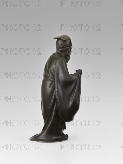 Sage, unknown sculptor, figurine, Chinese art, Chinese culture, unknown provenance; subject: sage man, scholar wearing a long flowing robe and a cap on his head; Qing dynasty 1644–1911, 19th century; material: sculpture; height 28.0 cm., A dark bronze statue of a robed figure shown in profile, standing with a long flowing garment that pools at the base and drapes over the arms; the figure wears a cap with a flat top and a decorative band at the back, has a beard and mustache, and one hand is extended forward while the other is held closer to the body; the surface shows smooth highlights and subtle wear, and the background is a light neutral gradient of gray and off-white.