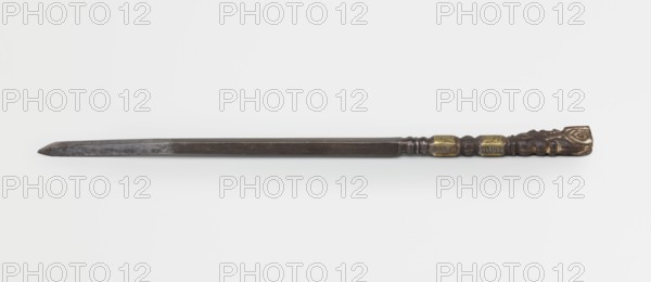 Dagger, 19th century, Tibetan culture, mililtaria weapon; daggers weapons; material: craft; origin: Tibet, height 25.8 cm, An elongated metal object with a long dark gray blade and a darker brownish handle; the handle features a carved end with a rounded decorative shape and a few raised bulbous sections, and includes a couple of small rectangular fittings with a golden-yellow tone set into the handle; the blade is straight with a tapered point and shows variations in darker and lighter gray, and the overall image background is a neutral pale off-white.