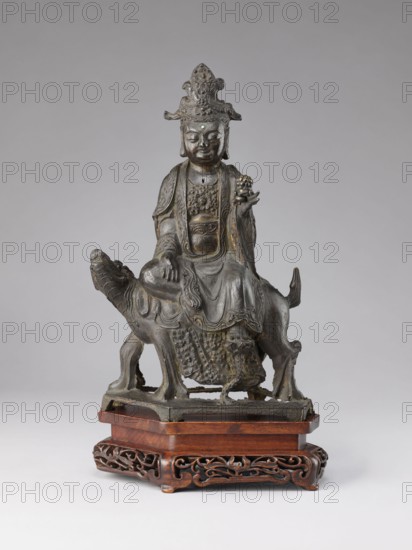 Buddhist deity on a buffalo, on a base; figurine; Chinese; Chinese Buddhism; lotus iconography; Qing dynasty 1644–1911, 19th century; sculpture; height 35.5 cm, width 21.5 cm;, A dark metal statue of a seated figure wearing an ornate headdress and flowing robes sits atop a stylized animal with a long tail, both resting on a carved wooden base; the figure holds a small rounded object in one hand and has detailed facial features and layered decorative elements on the clothing, while the metal shows areas of darker and slightly lighter patina and the wooden base is a warm reddish-brown with intricate openwork carving.