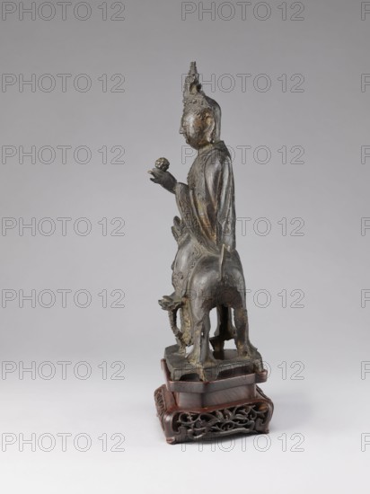 Buddhist deity on a buffalo on a base; figurine; Chinese; lotus iconography; Chinese Buddhism; Qing dynasty 1644–1911, 19th century; sculpture; height 35.5 cm, width 21.5 cm; maker, materials not specified., A bronze-colored sculptural figure with a dark patina stands on a carved wooden base; the figure wears a tall ornate headdress and flowing draped garments with visible folds and surface decoration, holding a small rounded object in one hand while the other hand rests near the body, and the figure is mounted on the back of an animal-like form with a short tail and sturdy legs. The wooden base is reddish-brown with an intricately carved openwork design and a tiered profile. The background is a smooth gradient of light gray, and the overall color palette includes bronze, dark gray, black, and reddish-brown.