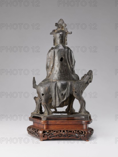 Buddhist deity on a buffalo on a base; bronze sculpture, Chinese, Qing 1644–1911, 19th century; lotus iconography; height 35.5 cm, width 21.5 cm., A dark metallic figurine of a seated person wearing a draped garment and a tall ornate headdress sits on the back of a sculpted animal with a raised head; the figure and animal show surface irregularities and worn areas, with several small openings visible in the metal; the sculpture rests on a carved wooden stand that is warm brown with intricate openwork scroll patterns; visible colors include dark gray, black, brown, and hints of lighter worn metal.