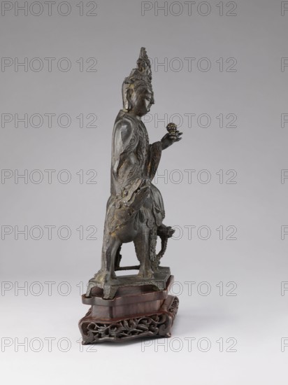 Buddhist deity on a buffalo on a base; figurine; Chinese; Chinese Buddhism; lotus iconography; Qing dynasty 1644–1911, 19th century; sculpture; height 35.5 cm, width 21.5 cm., A dark bronze sculpture of a standing figure wearing a tall ornate headdress and flowing robes stands on a carved wooden base; the figure faces sideways and holds a small rounded object in an outstretched hand, with detailed drapery and textured surfaces on the body and garment. The sculpture shows a few areas of lighter bronze or golden wear against the predominantly dark brown and black metal. The wooden base is a reddish brown with an intricately carved lower tier in a darker brown, and the background is a smooth neutral gray.