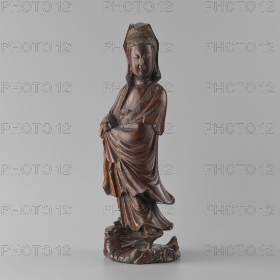 Guanyin Bodhisattva;, Qing dynasty 1644–1911, 19th century; China; sculpture; material: not specified; dimensions: height 31.6 cm, width 11.5 cm, depth 9.5 cm; subject: Guanyin Bodhisattva Chinese Buddhism, goddess/figurine; culture: Chinese., A carved wooden figurine of a standing robed figure with a smooth dark brown polished surface, wearing a headdress and layered flowing garments with carved folds and drapery, hands clasped in front holding a small rounded object, a serene facial expression with closed eyes and subtle features, the robe falling over a textured carved base that resembles rocks, all set against a plain light gray background with overall colors of dark brown, lighter brown highlights, and gray.