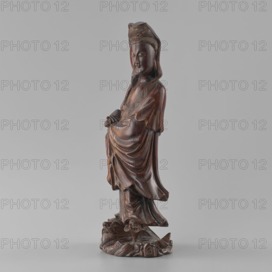 Guanyin Bodhisattva, sculptor unknown, Chinese art, figurine, Chinese goddess, Chinese Buddhism, Qing dynasty 1644–1911, 19th century, China; sculpture; height 31.6 cm, width 11.5 cm, depth 9.5 cm, A carved wooden figure of a robed person standing on an ornate base, with flowing drapery and folded hands, rendered in dark brown tones with variations of lighter brown highlights, set against a plain light gray background.