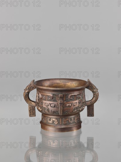 Gui-type incense burner, bronze, Chinese, Qing dynasty 1644–1911, 19th century. Ritual vessel gui with taotie and fantastical beasts iconography, height 10.0 cm, width 11.5 cm. Creator,  noted., An ornate metal vessel with a rounded bowl and a short cylindrical base sits on a reflective surface, showing a mirrored reflection beneath it; the vessel is bronze in color with areas of darker brown and hints of greenish patina, and it features a pair of curved handles with rectangular hanging plaques and an all-over carved pattern of stylized animal and geometric motifs wrapping around the body and base.