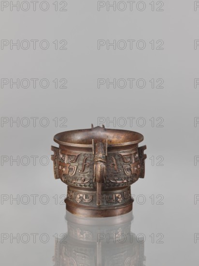 Gui-type incense burner, bronze, Chinese, Qing dynasty 1644–1911, 19th century, China; gui vessel with taotie mask and fantastical beasts, ritual/ceremonial vessel; height 10.0 cm, width 11.5 cm., A bronze vessel with a rounded rim and a slightly flared mouth sits centered against a neutral gray background, showing a warm brown and dark brown patina with subtle greenish and reddish tints; the vessel has a short cylindrical foot and a wider body decorated with raised geometric and zoomorphic reliefs forming repeating patterns around the exterior, and a vertical bronze strap or handle running from rim to base with a stylized animal head at its top; a faint reflection of the vessel appears on the glossy surface beneath it.