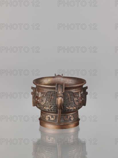Gui-type incense burner, bronze, Chinese, Qing dynasty 1644–1911, 19th century, gui vessel with taotie monster motif and fantastical beasts, ritual/ceremonial vessel, height 10.0 cm, width 11.5 cm., A metal vessel with a rounded bowl and a short cylindrical base sits centered on a reflective surface, showing a faint mirrored image beneath; the vessel is brown with darker and lighter bronze tones and has intricate raised geometric and stylized motifs carved around its body, including decorative protrusions and a vertical central element on the front; the rim is slightly flared and there are small tab-like extensions on the sides, while the background is a uniform light gray.
