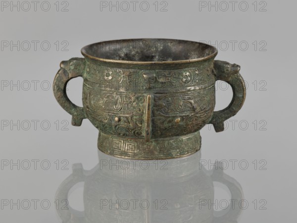 Gui incense burner, Chinese, ceremonial ritual vessel gui, 19th century Qing 1644–1911, taotie and meander leiwen ornamentation, material unspecified, height 6.5 cm, width 12.8 cm, diameter 8.9 cm., An aged metal vessel with a rounded bowl and a short flared rim sits centered on a reflective surface, showing a mirrored image beneath; the vessel has two looped handles on opposite sides and a short cylindrical base, all covered in intricate raised decorative patterns and motifs in a worn, textured finish. The visible colors include greenish patina, dark brown, muted bronze, and hints of black, with areas of lighter metallic sheen where the surface is more exposed. The interior appears darker and mottled, the surface shows irregular corrosion and verdigris, and the reflections on the base echo the vessel’s shape and colors.