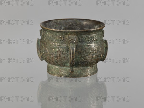 Gui incense burner, Chinese, ceremonial vessel with taotie and meander ornament, leiwen ornament, ritual gui vessel, Qing dynasty 1644–1911, 19th century, China; height 6.5 cm, width 12.8 cm, diameter 8.9 cm, A small ornate metal vessel sits centered on a reflective surface, showing a greenish patina with areas of darker brown and hints of bluish corrosion; the vessel has a rounded body with a wide open rim, decorative raised patterns all around its exterior, and a few short strap-like handles projecting from the sides and front; the interior appears darker and worn, and the reflection beneath repeats the vessel's shape and colors on a pale gray background.