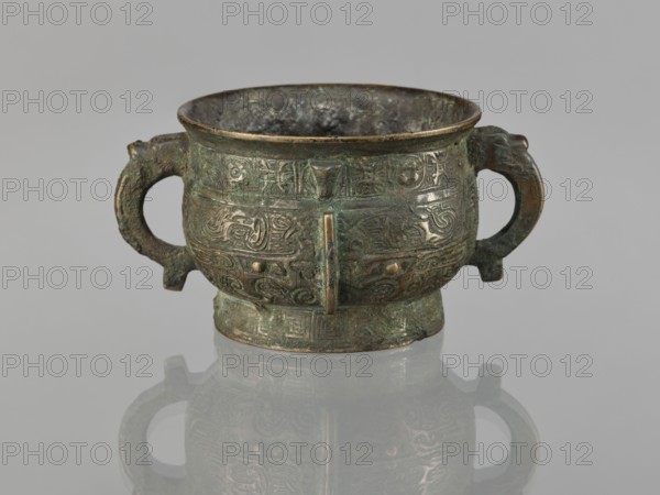 Gui incense burner, Chinese, ceremonial vessel, taotie iconography, meander and leiwen ornament, ritual vessel, Qing dynasty 1644–1911, 19th century, China, vessel; height 6.5 cm, width 12.8 cm, diameter 8.9 cm, An aged metal vessel with a rounded body and a short pedestal base sits centered on a reflective surface, showing a faint mirror image beneath it; the vessel has two looped handles on opposite sides and a wide rim, and its surface is covered in intricate raised patterns and textured motifs; the metal shows a patina with green and brown tones alongside darker areas of tarnish, and the reflection echoes those green, brown, and dark hues against a pale gray background.