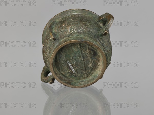 Gui incense burner, Chinese, ceremonial vessel with taotie and meander ornamentation, leiwen pattern, Qing dynasty 1644–1911, 19th century, China, vessel gui, height 6.5 cm, width 12.8 cm, diameter 8.9 cm, An old metal vessel photographed from an angled top view, showing ornate raised patterns on its exterior surface and a rounded rim; the vessel has a greenish patina with areas of brown and darker metal tones, and two small looped handles on opposite sides; the interior shows textured corrosion in green and brown hues and the vessel casts a soft reflection on a pale gray surface.