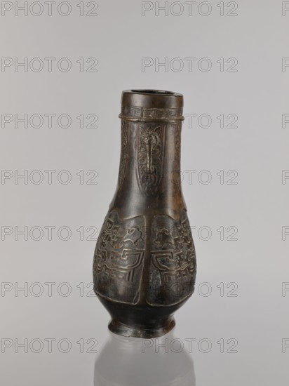 Hu-type vessel, bronzes, Chinese art, Chinese culture, Qing 1644–1911, ceremonial/ritual vessel, taotie iconography, meander ornament, Qing style, 19th century, China, vessel, height 28.0 cm, width 19.0 cm, A carved vase-like object stands upright on a reflective surface, showing a dark brown to nearly black patina with lighter brown highlights on raised areas; the object tapers from a wider rounded midsection to a narrower neck and flared rim, and the body features repeated carved panels with stylized animal or abstract motifs and patterned borders, while the neck displays elongated vertical decorative panels and a band of geometric ornament near the rim; the base is slightly flared and the overall background is a soft gray.